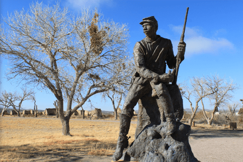 A bronze statue of a soldier holding a rifle, set against a backdrop of bare trees and a blue sky.