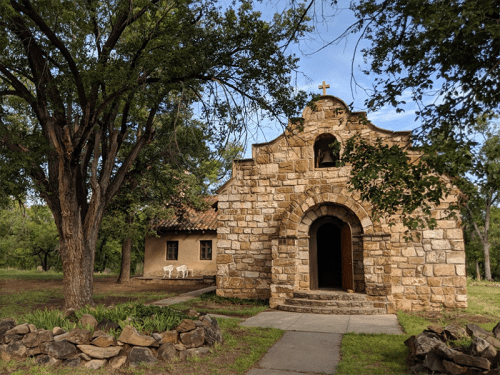 A rustic stone chapel with a cross, surrounded by trees and a small building in a serene outdoor setting.