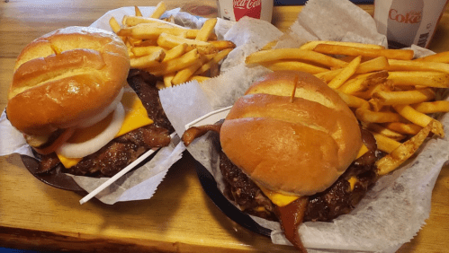 Two burgers with cheese, bacon, and toppings, served with a side of golden French fries on a wooden table.