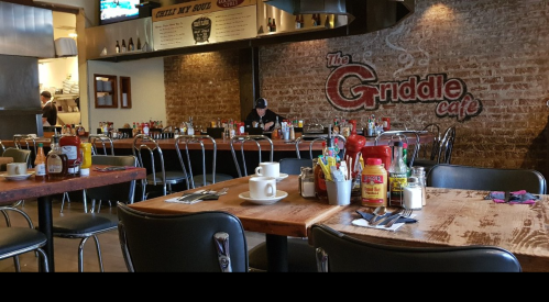 Interior of The Griddle Cafe with wooden tables, chairs, and a wall featuring the cafe's name. Condiments are on the tables.