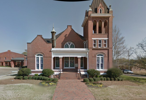 Historic brick building with a tall tower, arched windows, and landscaped front yard.