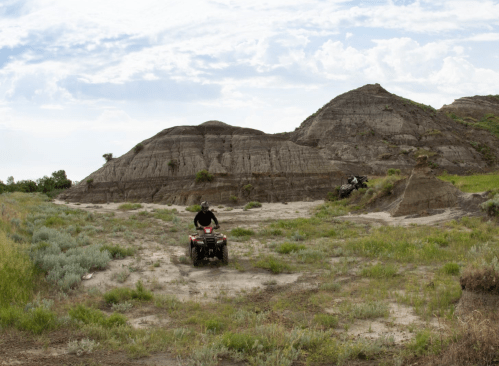 A person rides an ATV through a rugged landscape with hills and sparse vegetation under a cloudy sky.