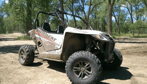 A muddy all-terrain vehicle parked on a dirt path surrounded by trees and blue sky.