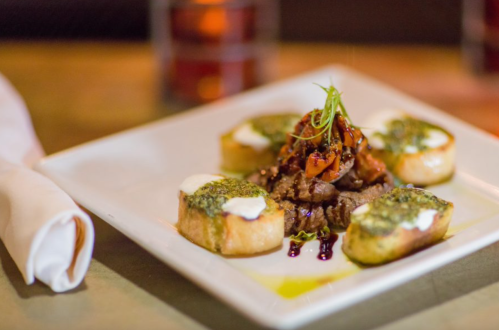 A gourmet dish featuring grilled meat topped with herbs, served with pesto-covered bread and a drizzle of sauce.