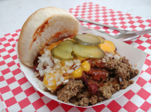 A hearty sandwich with ground meat, cheese, pickles, and rice, served on a bun, on a red and white checkered tablecloth.