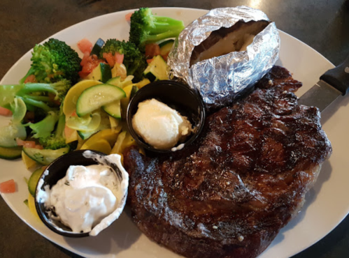 A plate featuring a grilled steak, baked potato, butter, and a side of mixed vegetables including broccoli and zucchini.