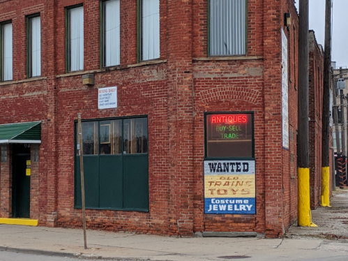 A brick building with a neon sign reading "Antiques Buy-Sell-Trade" and a "Wanted" sign for old trains and costume jewelry.