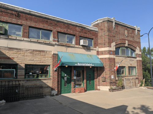 Historic brick building with green doors and awning, featuring flags and large windows under a clear blue sky.