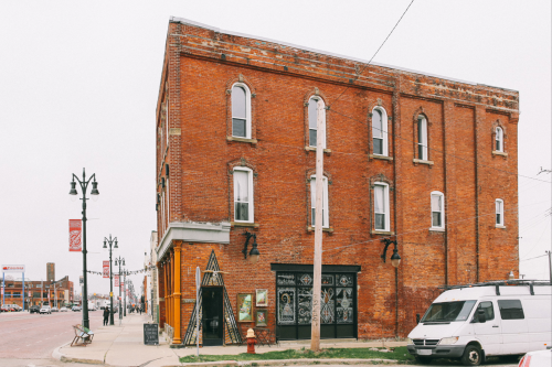 A brick building with large windows, featuring a wooden entrance and a parked van nearby on a cloudy day.