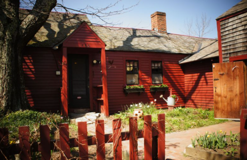 A red wooden house with a sloped roof, surrounded by greenery and a wooden fence, under a clear blue sky.