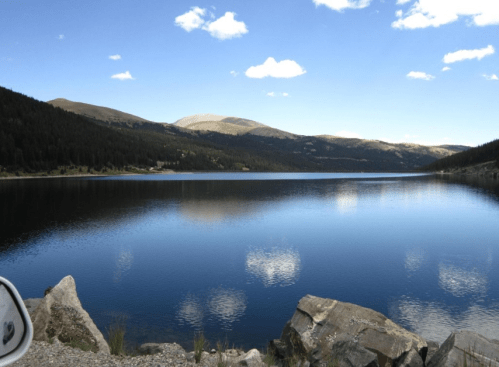 A serene lake surrounded by mountains and trees, reflecting clouds in the clear blue sky.
