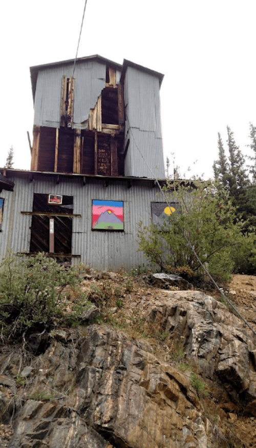 A dilapidated metal building on a rocky hillside, featuring colorful paintings on its walls and a partially collapsed roof.