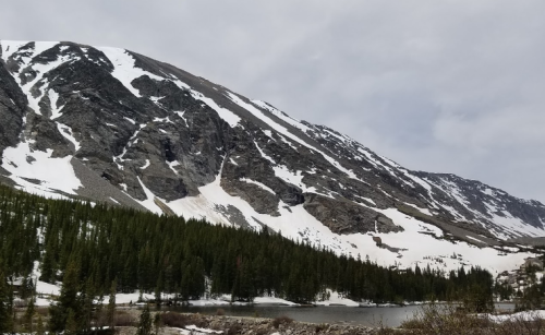 Snow-capped mountains rise above a forested area beside a calm lake under a cloudy sky.