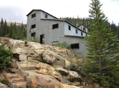 Abandoned building on rocky terrain, surrounded by trees and mountains under a cloudy sky.