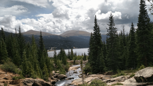 A scenic view of a lake surrounded by mountains and dense evergreen trees under a cloudy sky.