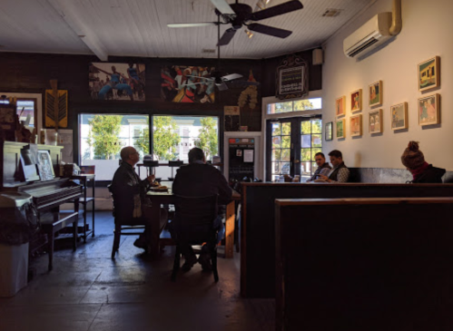 A cozy café interior with patrons seated at tables, artwork on the walls, and large windows letting in natural light.