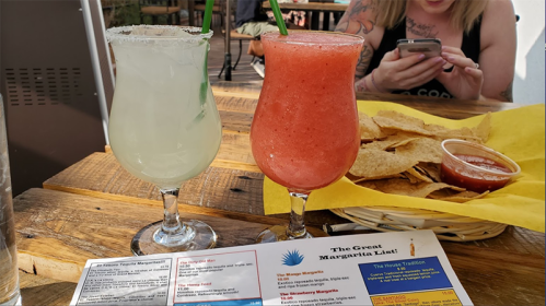 Two colorful cocktails on a table, one green and one pink, with a basket of tortilla chips and salsa in the background.