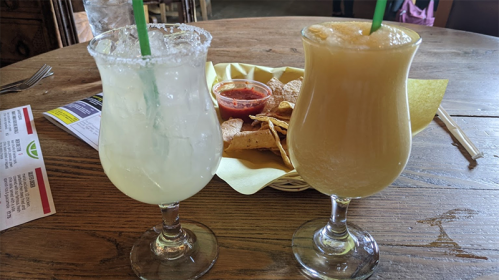 Two colorful cocktails with straws sit on a wooden table, accompanied by a basket of tortilla chips and salsa.