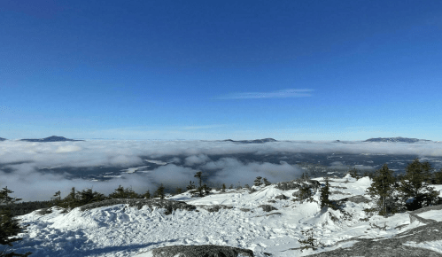 A snowy mountain landscape under a clear blue sky, with clouds rolling over the valleys below.