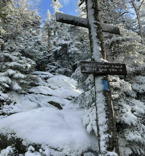 Snow-covered trail sign in a winter forest, indicating directions to Mount Hunger and White Peak.