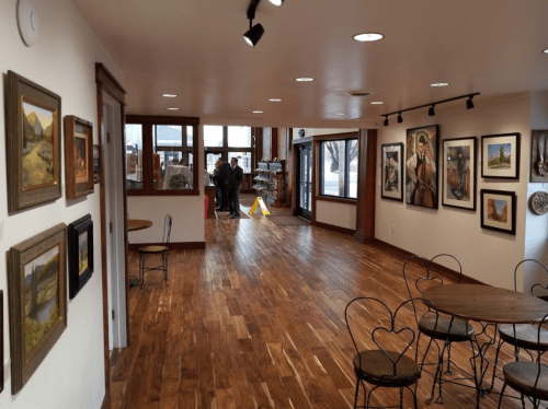 A spacious gallery with wooden floors, featuring artwork on the walls and tables, with people in the background.