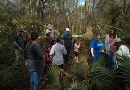 A group of people, including children, gathered in a wooded area, surrounded by greenery and trees.