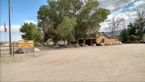 A rustic building named "The Boonies" surrounded by trees and large rocks, set in a dusty outdoor area.