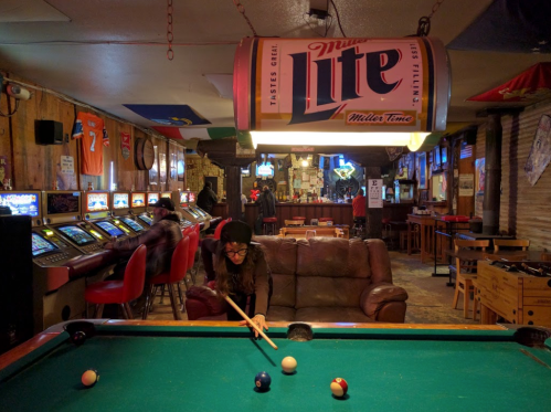 A woman plays pool in a cozy bar with a large Miller Lite sign overhead and various gaming machines in the background.
