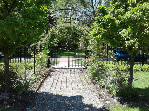 A garden archway surrounded by trees, leading to a pathway and a view of a street beyond.