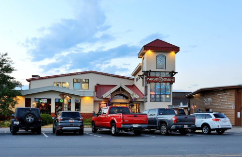A restaurant with a clock tower and outdoor seating, surrounded by parked cars at dusk.