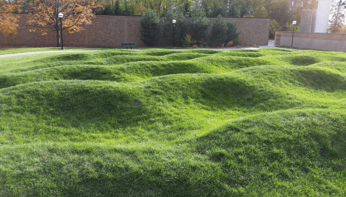 A grassy landscape with rolling hills and gentle mounds, surrounded by trees and a brick wall in the background.