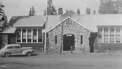 Historic stone building with large windows, a flagpole, and vintage cars parked outside, surrounded by trees.