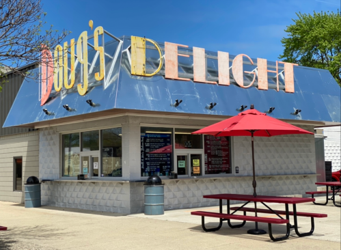 A colorful food stand named "Doug's Delight" with a red umbrella and picnic table outside on a sunny day.