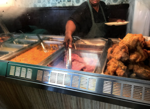 A server in an apron uses tongs to serve food from a buffet, featuring fried chicken and sides.