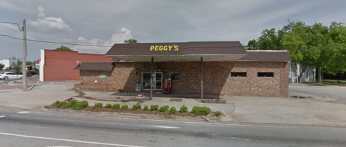 A brick building with a sign reading "Peggy's," featuring a soda machine outside and a clear blue sky.