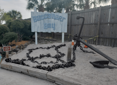 Sign for Buccaneer Bay with a large anchor and chain displayed in a sandy area, surrounded by greenery and a wooden fence.