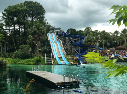 A water park scene featuring two blue water slides beside a clear, greenish-blue pool under a cloudy sky.