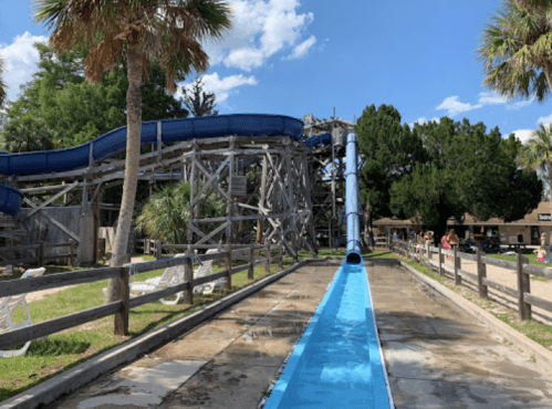 A water slide at a park, with a blue slide curving down and people enjoying the area under a clear blue sky.