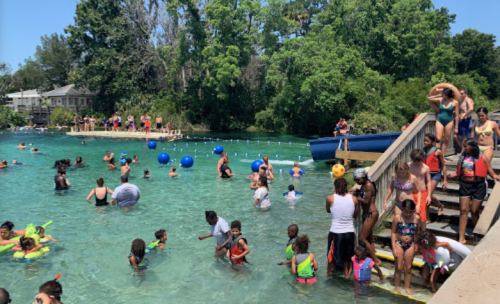 A lively swimming area with people of all ages enjoying the water, colorful floats, and a sunny day.
