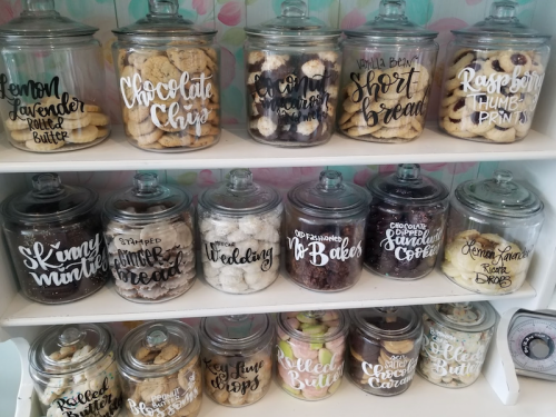 A display of labeled glass jars filled with various baked goods, including cookies and breads, on a shelf.