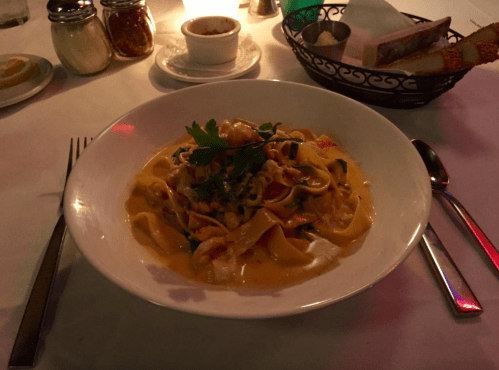 A bowl of creamy pasta with shrimp and herbs, set on a table with a candle and condiments in the background.