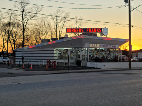 A diner named "Burgers Franks" with a neon sign, surrounded by trees at sunset.