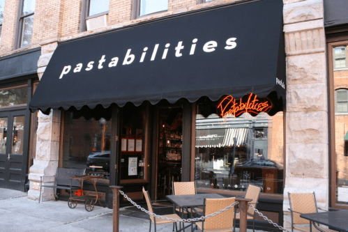 Exterior of a restaurant named "pastabilities," featuring a black awning and outdoor seating. Neon sign visible.
