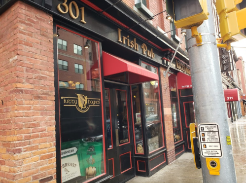 Exterior of an Irish pub with red awnings, brick walls, and a street corner view. Rainy weather is visible.