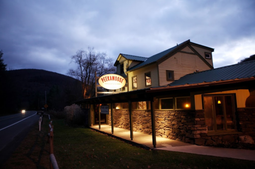 A cozy restaurant named Peekamoose, illuminated at dusk, with a stone facade and mountains in the background.