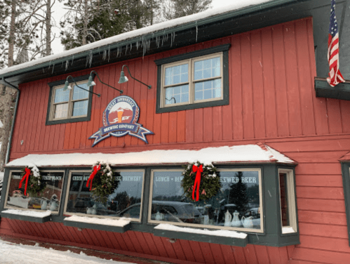 A red wooden building with wreaths on the windows, featuring a sign for a brewery, surrounded by snow and trees.