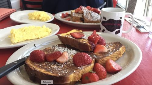 A plate of French toast topped with strawberries, alongside scrambled eggs and a coffee mug on a red table.