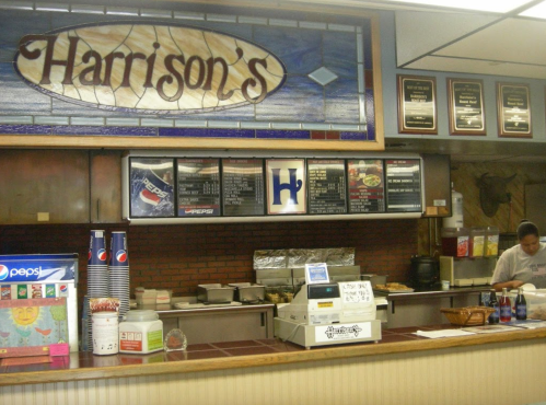 Interior of Harrison's restaurant featuring a menu board, cash register, and a staff member preparing food.