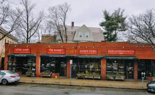 A row of brick storefronts featuring a wine shop, market, and pasta shop, with trees and a residential building in the background.