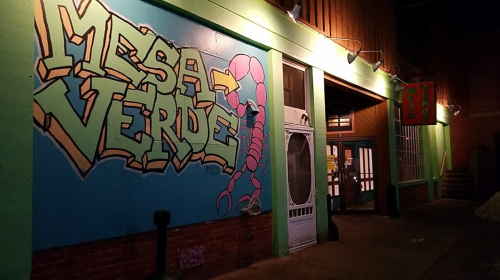 Colorful mural of "Mesa Verde" with a scorpion, outside a building at night. Brightly lit entrance visible.